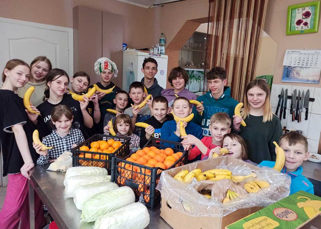 De kinderen van het kinderhuis staan in de keuken met kratten vers fruit. Ze hebben bananen in hun hand.